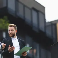 Dwóch agentów nieruchomości dyskutuje, jak wycenić mieszkanie, analizując dokumenty na tle nowoczesnego budynku.