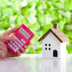 Różowa kalkulatorka w dłoniach obok modelu domu. Obrazek symbolizuje dofinansowanie do wynajmu mieszkania dla młodych.
