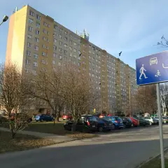 Długi blok mieszkalny, parking z samochodami i znak informujący o możliwości wykupu mieszkania spółdzielczego.