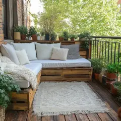 Przytulny balkon w bloku, gdzie drewniane palety tworzą sofę. Rośliny i dywanik dodają uroku, tworząc idealne miejsce do relaksu.