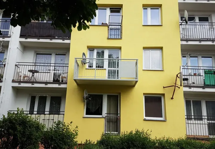Żółty blok z dobudową balkonu na słupach. Widać okna, balustrady i anteny satelitarne.