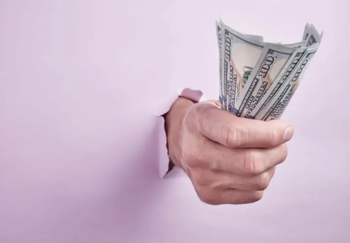 Dłoń przebija się przez różową ścianę, trzymając plik banknotów 100 USD. Ktoś walczy o zwrot kaucji.