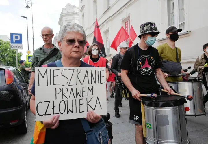 Kobieta trzyma transparent: "MIESZKANIE PRAWEM CZŁOWIEKA". W tle protestujący z bębnami i flagami.