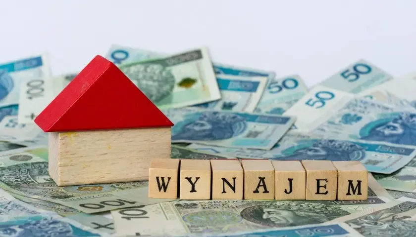 Drewniany domek i kostki z napisem "WYNAJEM" na tle polskich banknotów. Zgłoszenie umowy najmu do urzędu skarbowego.