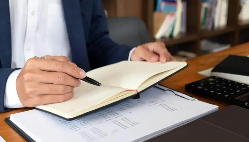 Mężczyzna w garniturze notuje w dzienniku, obok leży kalkulator i dokumenty z przykładowym numerem księgi wieczystej.