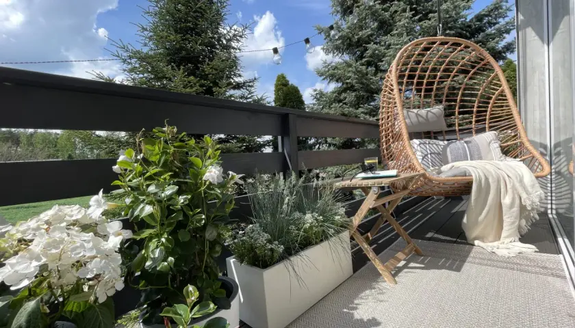 Przytulny mini balkon w bloku z fotelem wiszącym, stolikiem i kwitnącymi roślinami. Idealne miejsce na relaks.