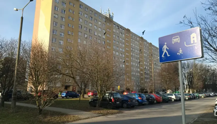 Długi blok mieszkalny, parking z samochodami i znak informujący o możliwości wykupu mieszkania spółdzielczego.