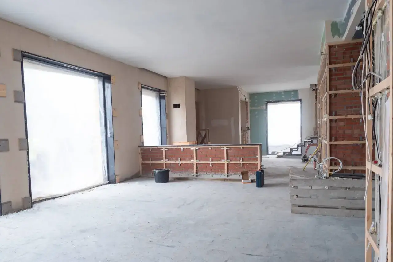 Wnętrze domu w stanie deweloperskim. Widać surowe ściany, instalacje, okna zabezpieczone folią i fragmenty ceglanej zabudowy.