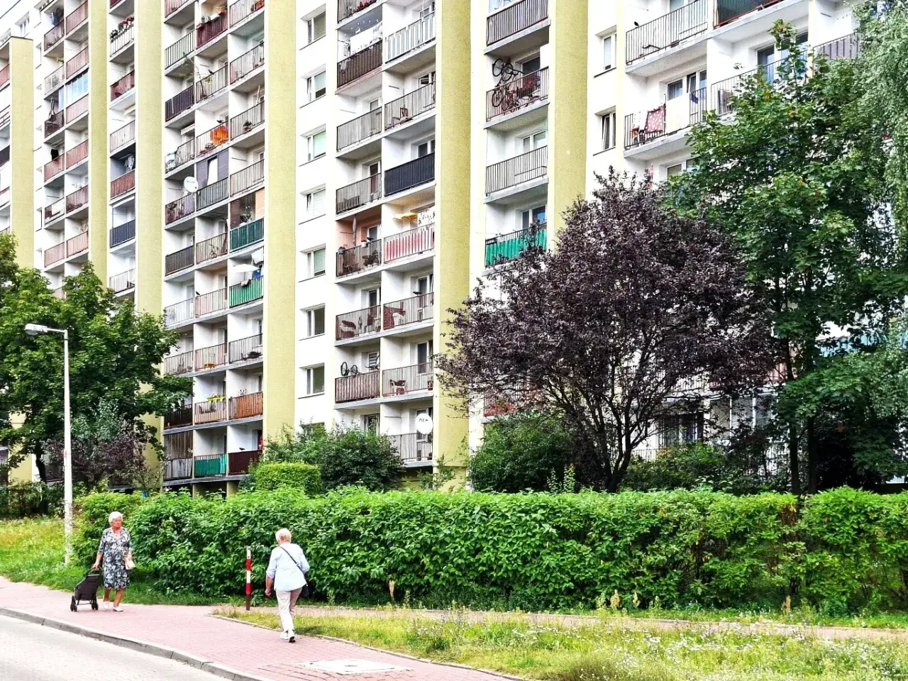Blok mieszkalny z balkonami, zielony żywopłot i spacerujące panie. W tle widać drzewa, a całość sugeruje zadbane osiedle, gdzie fundusz remontowy w spółdzielni mieszkaniowej dba o estetykę.