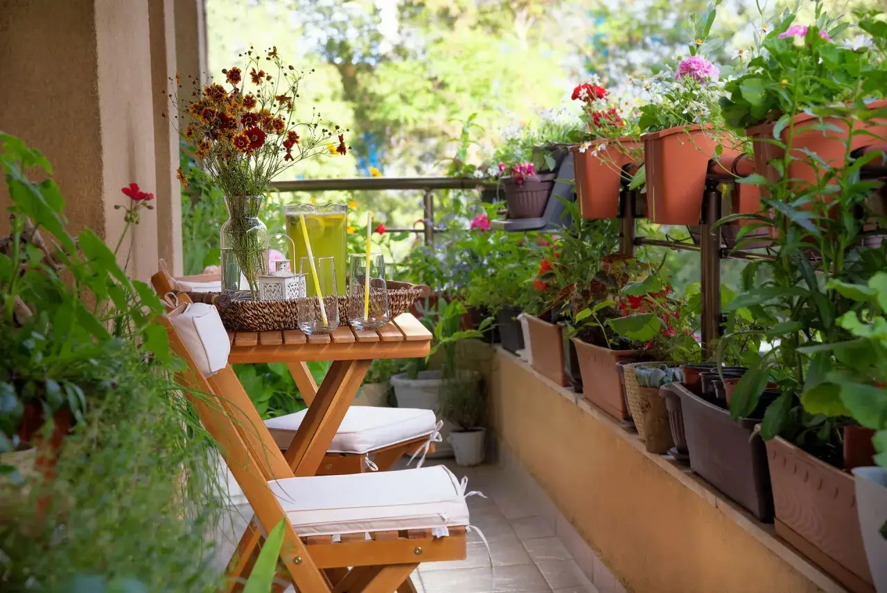 Uroczy mini balkon w bloku, pełen zieleni i kwiatów. Drewniany stolik z krzesłami, dzbanek z lemoniadą i bukiet kwiatów zapraszają do relaksu.