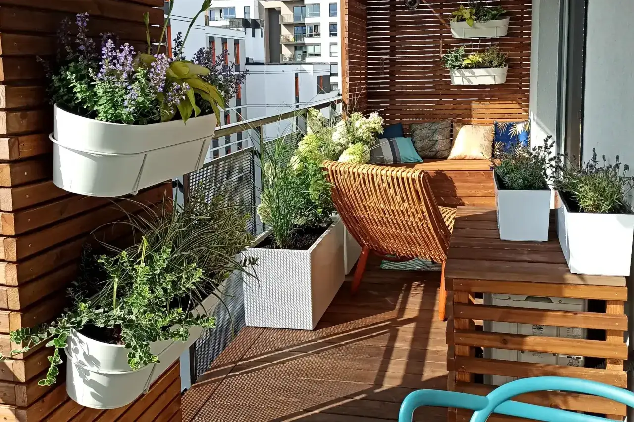 Zielony, przytulny mini balkon w bloku z rattanowym fotelem, ławką i mnóstwem kwiatów w białych donicach.