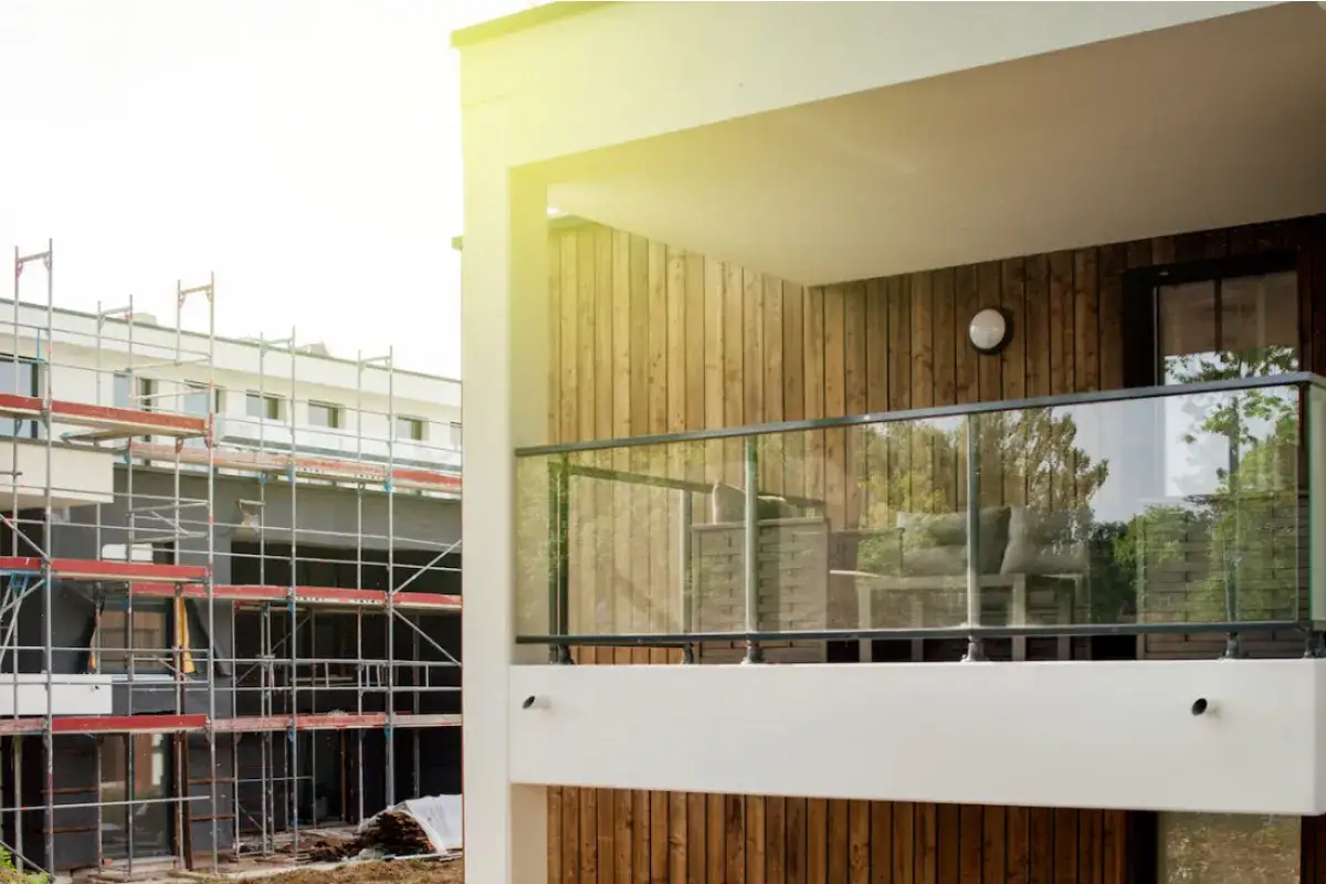 Nowoczesna loggia w bloku z przeszkloną balustradą i drewnianą elewacją. W tle budynek w budowie ze szkodami.