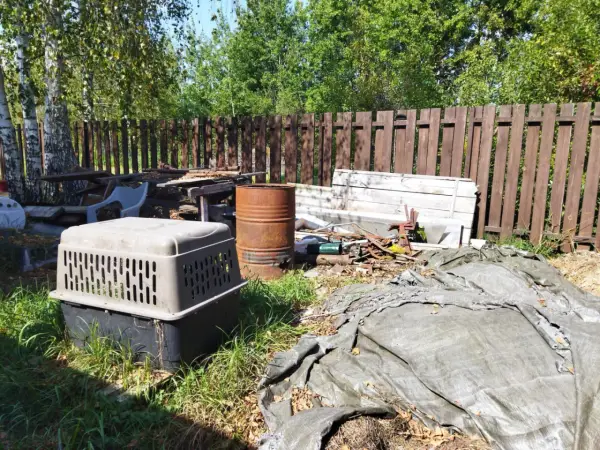 Zarośnięty ogród z rupieciami, beczką i klatką dla psa. Wygląda jak podwórko uciążliwego sąsiada na wsi.