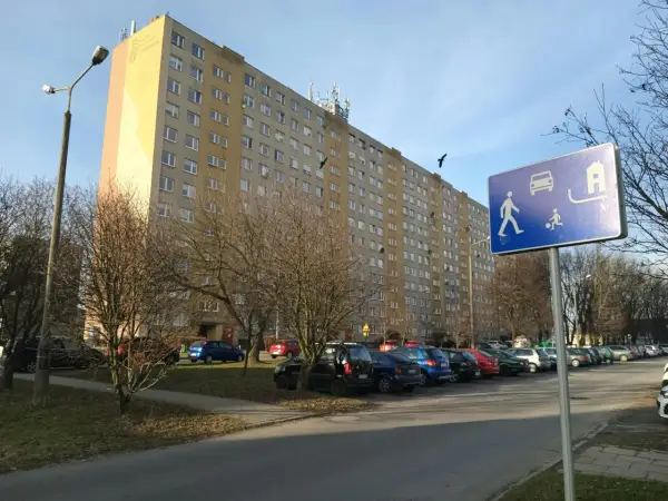 Długi blok mieszkalny, parking z samochodami i znak informujący o możliwości wykupu mieszkania spółdzielczego.